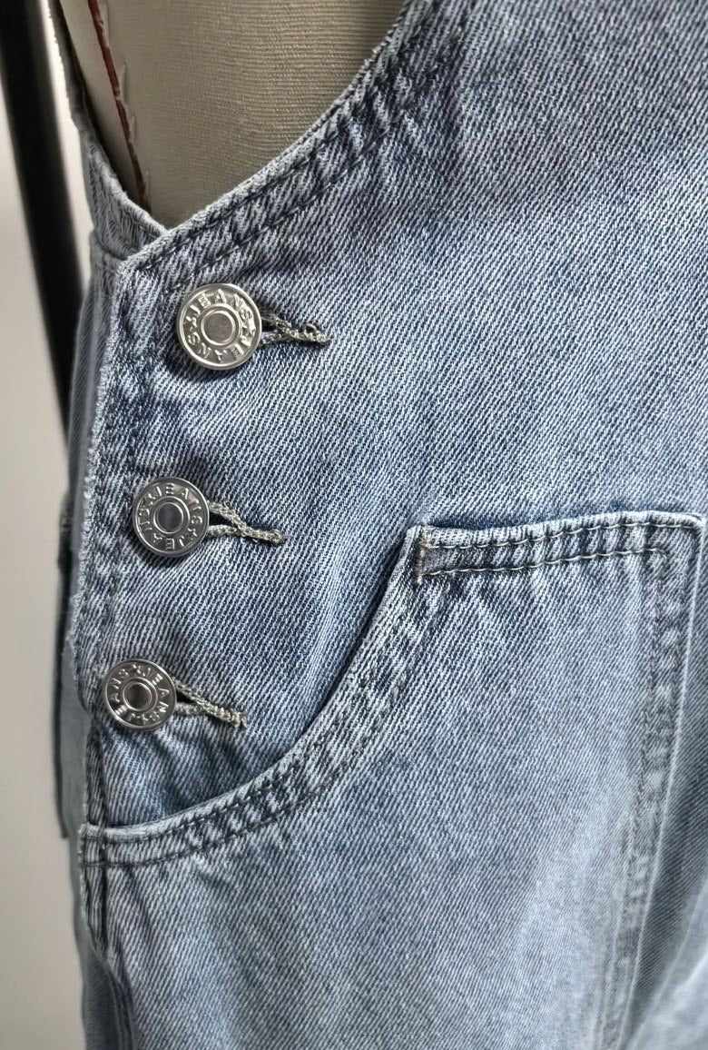 Close-up of metal side buttons and front patch pocket on women’s light blue denim overalls